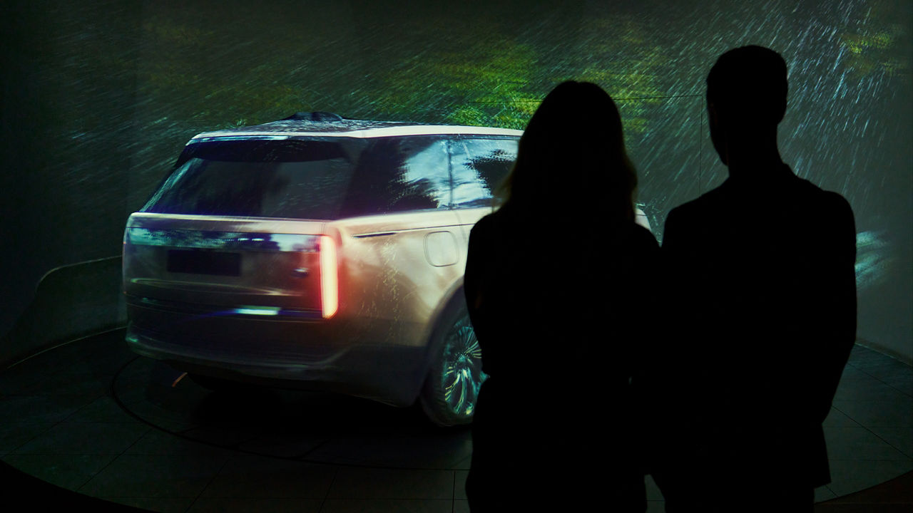 Silhouetted guests viewing an immersive 3D projection mapping reveal of a Range Rover, featuring dynamic lighting and surround‑sound effects that bring the vehicle’s design and character to life as part of the Range Rover Studio’s immersive collection experience.