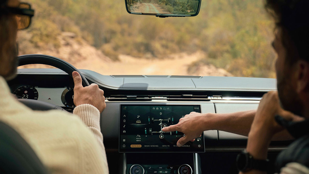 Two people inside a Range Rover reviewing off‑road driving settings on the touchscreen display while navigating a rugged trail during a Land Rover Experience activity.