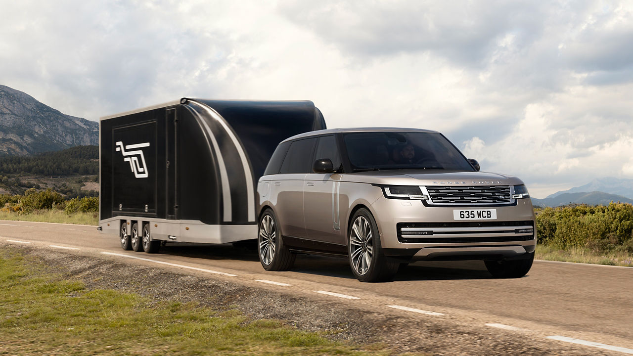 Range Rover towing a large, aerodynamic black enclosed trailer along a country road against a mountain backdrop.
